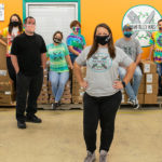 11 people stand in a brightly-colored kitchen while wearing masks