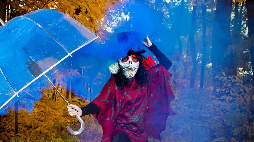 Woman wearing skeleton mask holding an umbrella in blue smoke.