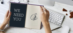 hands holding a book with a keyboard in the shot, book is open to a page that says WE NEED YOU with a finger pointing to the reader