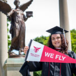 Kaitlynn Holmes holding a "We Fly" pennant in front of the Beneficence statue