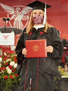 Office of Disability Services' Assistant Director, Jordan Grammas, pictured at Ball State University's masters' programs commencement ceremony. Jordan is wearing a black master's robe, hood, and cap. She is holding her red diploma case on the commencement stage.