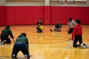 Six people are spread out across a basketball court. They are blindfolded and on their hands and knees. One person on the far side of the court holds a yellow ball.